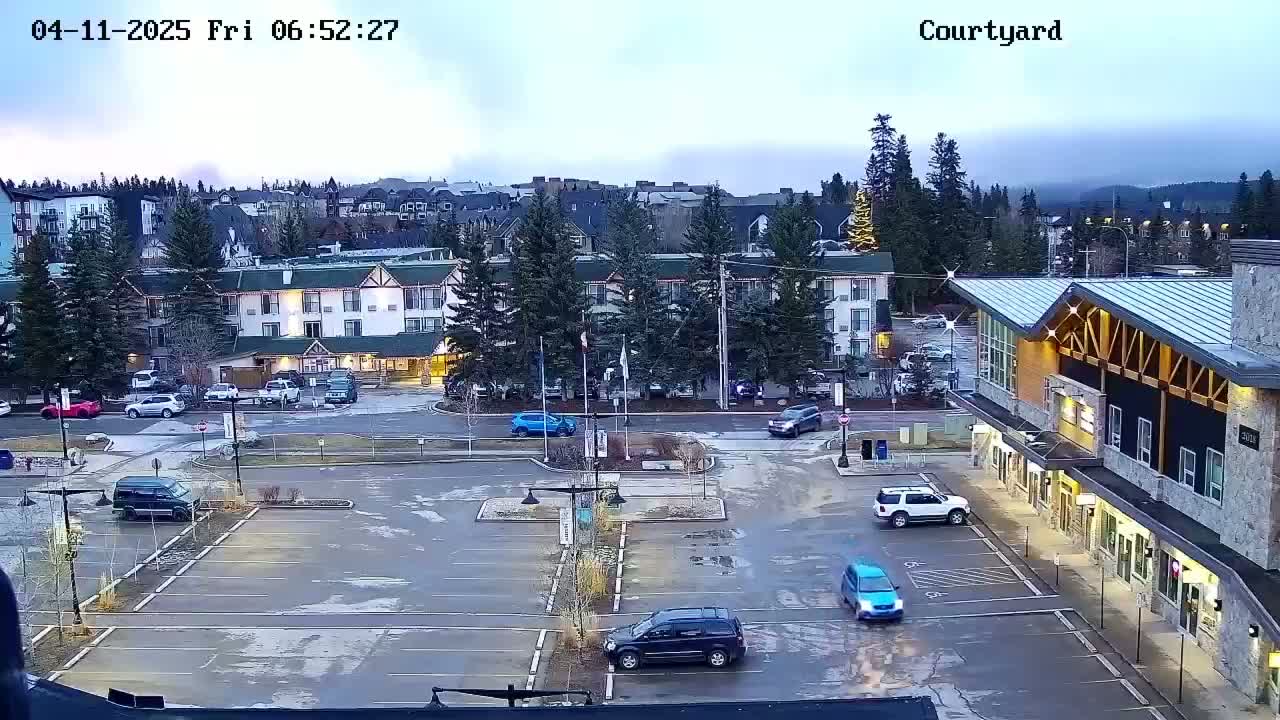 A mostly empty parking lot with several cars, a hotel, and other buildings under an overcast sky.