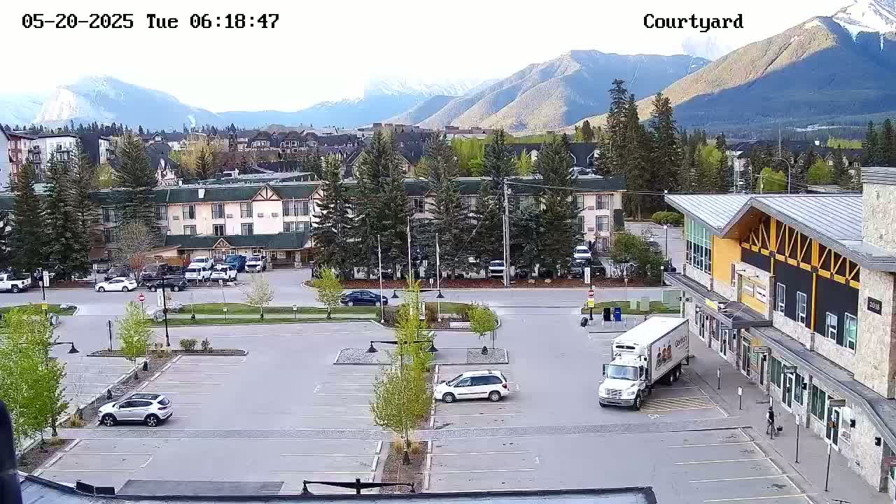 A parking lot with several cars and a delivery truck is shown, next to a building in front of a hotel with mountains in the background under a clear sky.