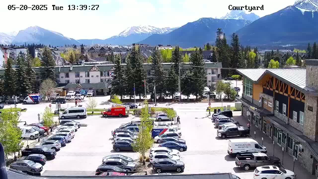 A sunny parking lot with numerous cars is situated in front of a multi-story building and other structures, all set against a backdrop of snow-capped mountains.