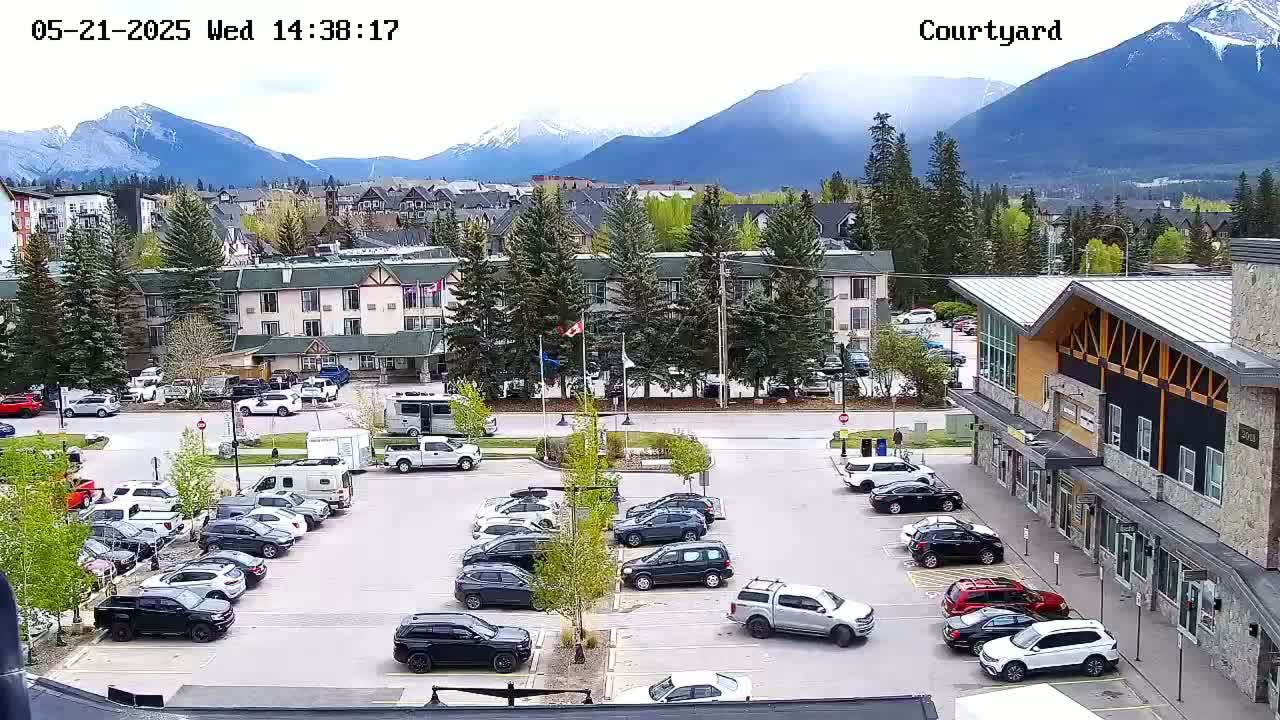 A parking lot filled with cars is situated in front of several buildings, with a mountain range in the background under partly cloudy skies.