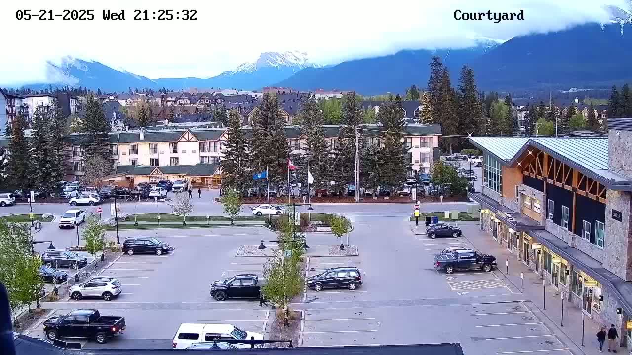 A parking lot with numerous vehicles is situated in front of a multi-story building, with a mountain range visible in the background under a partly cloudy sky.