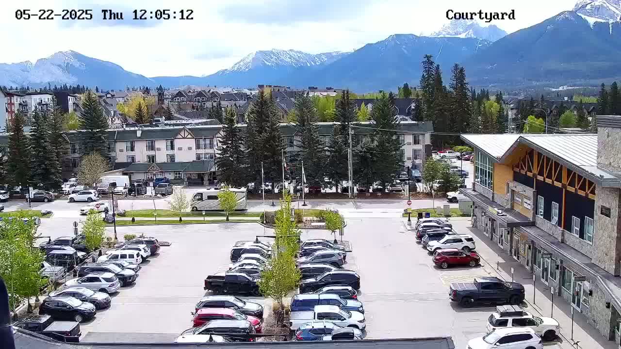A parking lot filled with cars is situated in front of buildings, with a mountain range in the background under a partly cloudy sky.