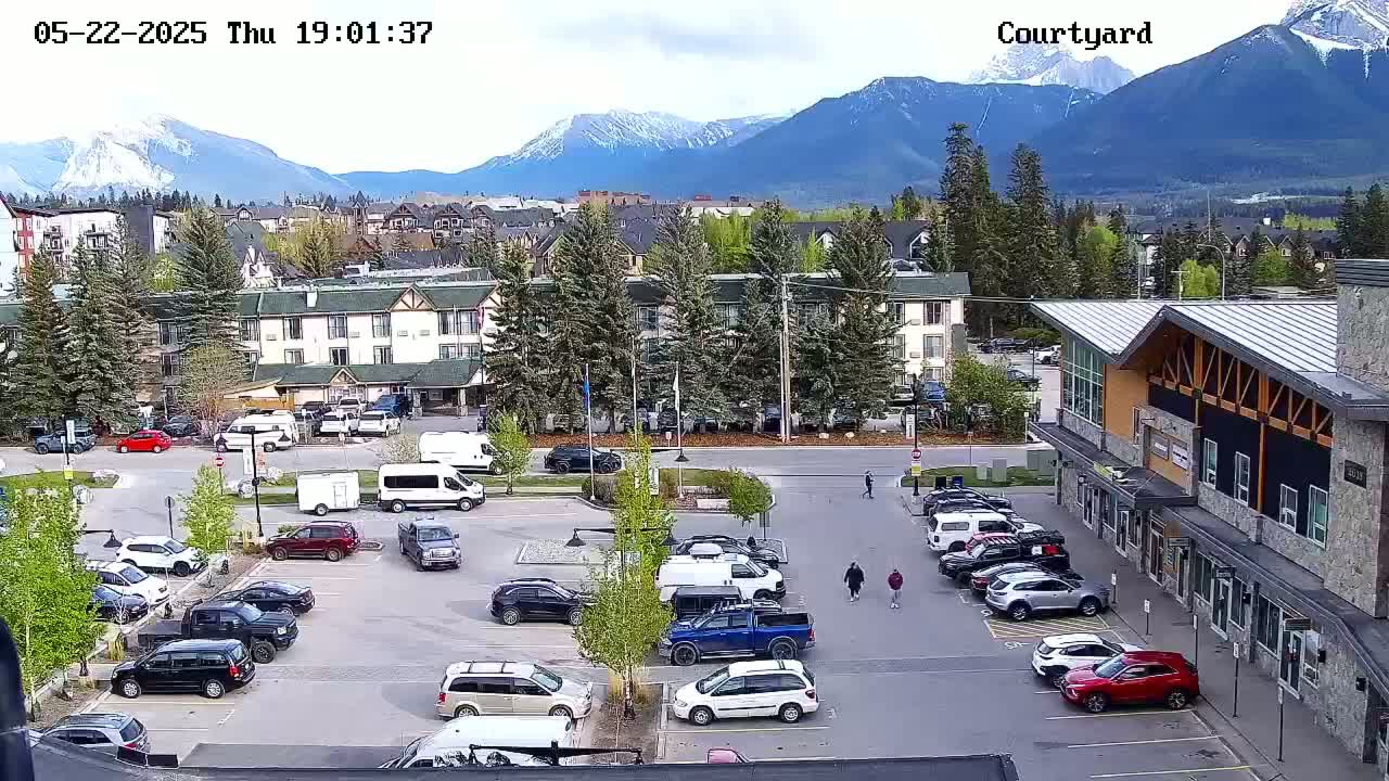 A parking lot with numerous cars and a hotel in the background, situated in front of a mountain range under a partly cloudy sky.