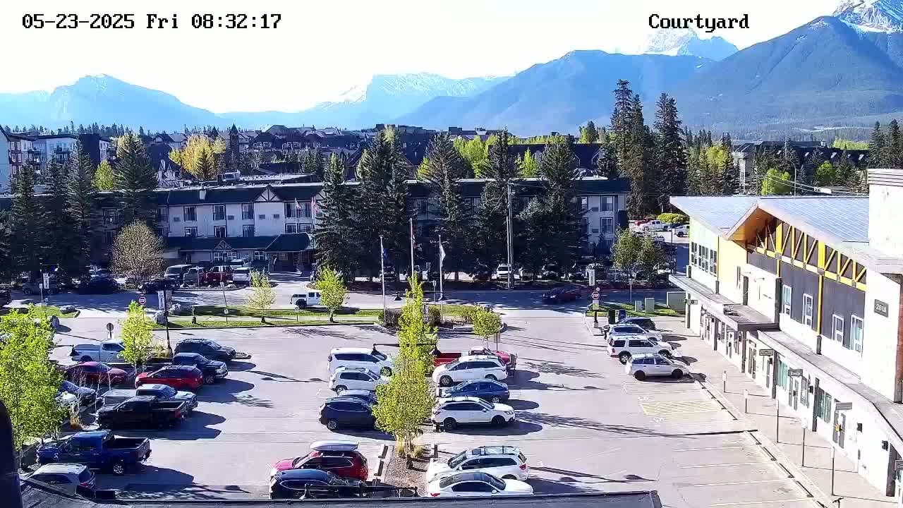 A parking lot with numerous cars is situated in front of buildings, all set against a backdrop of mountains under a clear, sunny sky.
