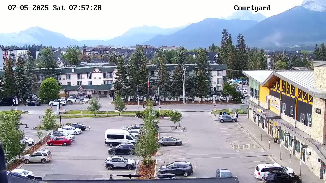 A parking lot with cars, a multi-story hotel, and other buildings is situated in front of a mountain range under a partly cloudy sky.