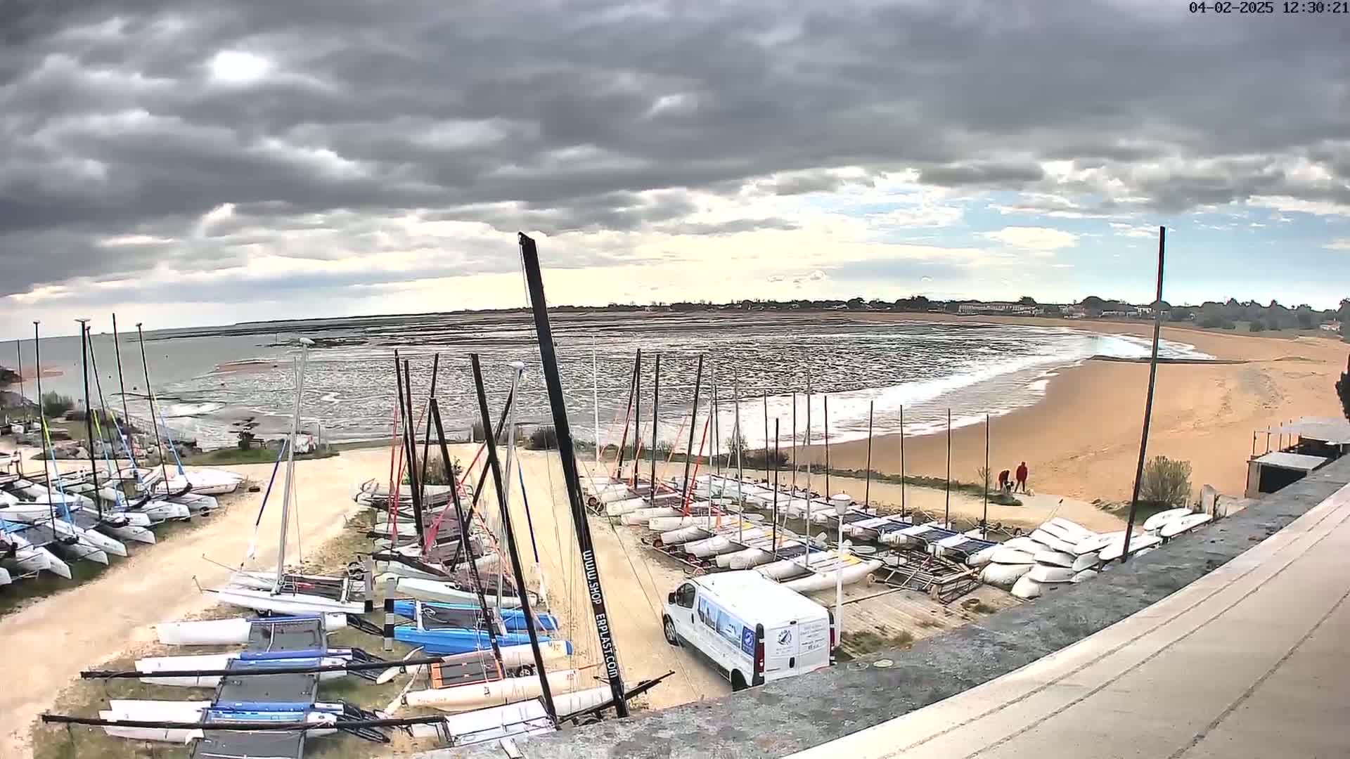 Numerous sailboats are stored on the ground near a sandy beach with small waves under a cloudy sky.