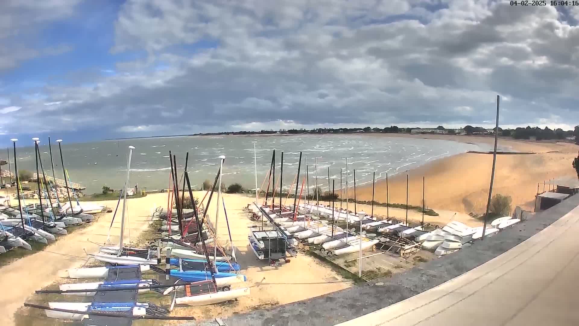 Many sailboats are stored on a sandy shore next to a body of water with choppy waves under a partly cloudy sky.