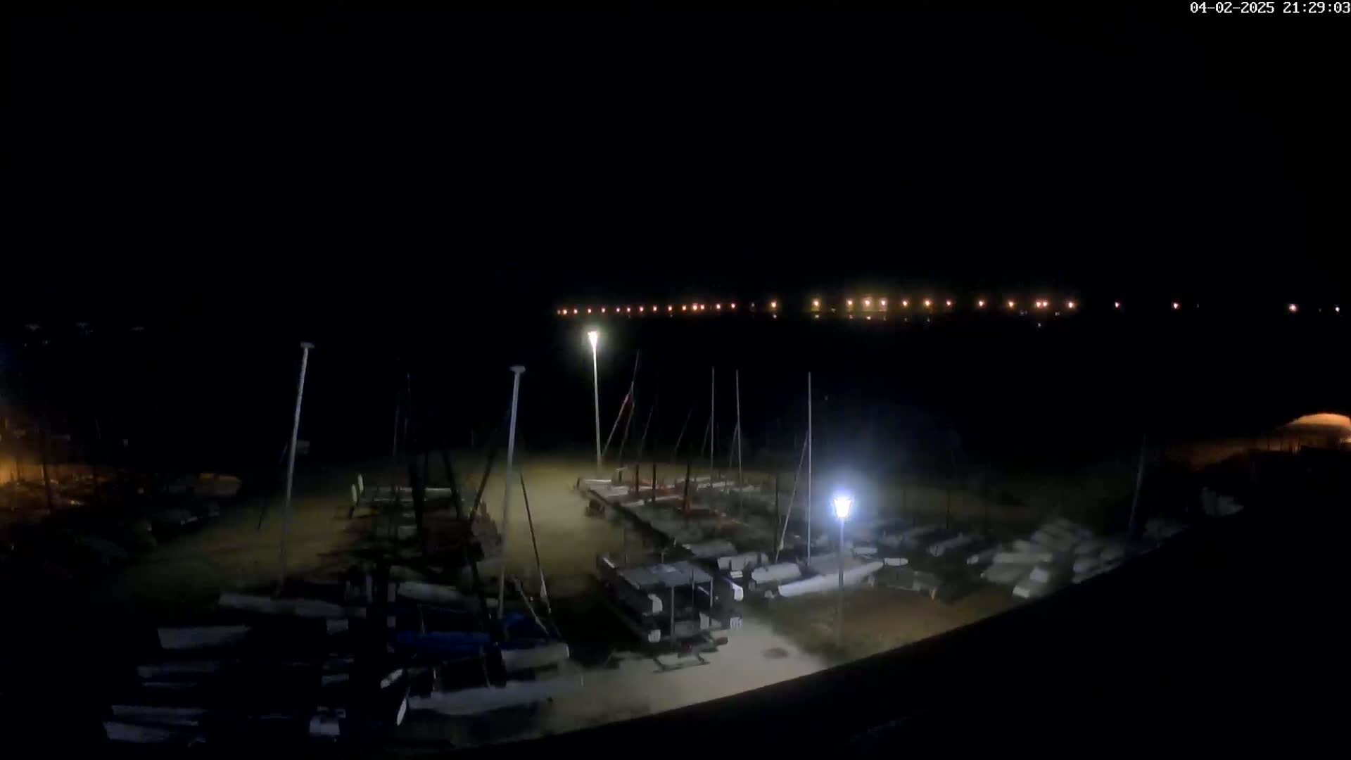 At night, numerous sailboats are docked in a marina, with a distant line of lights visible across the water.