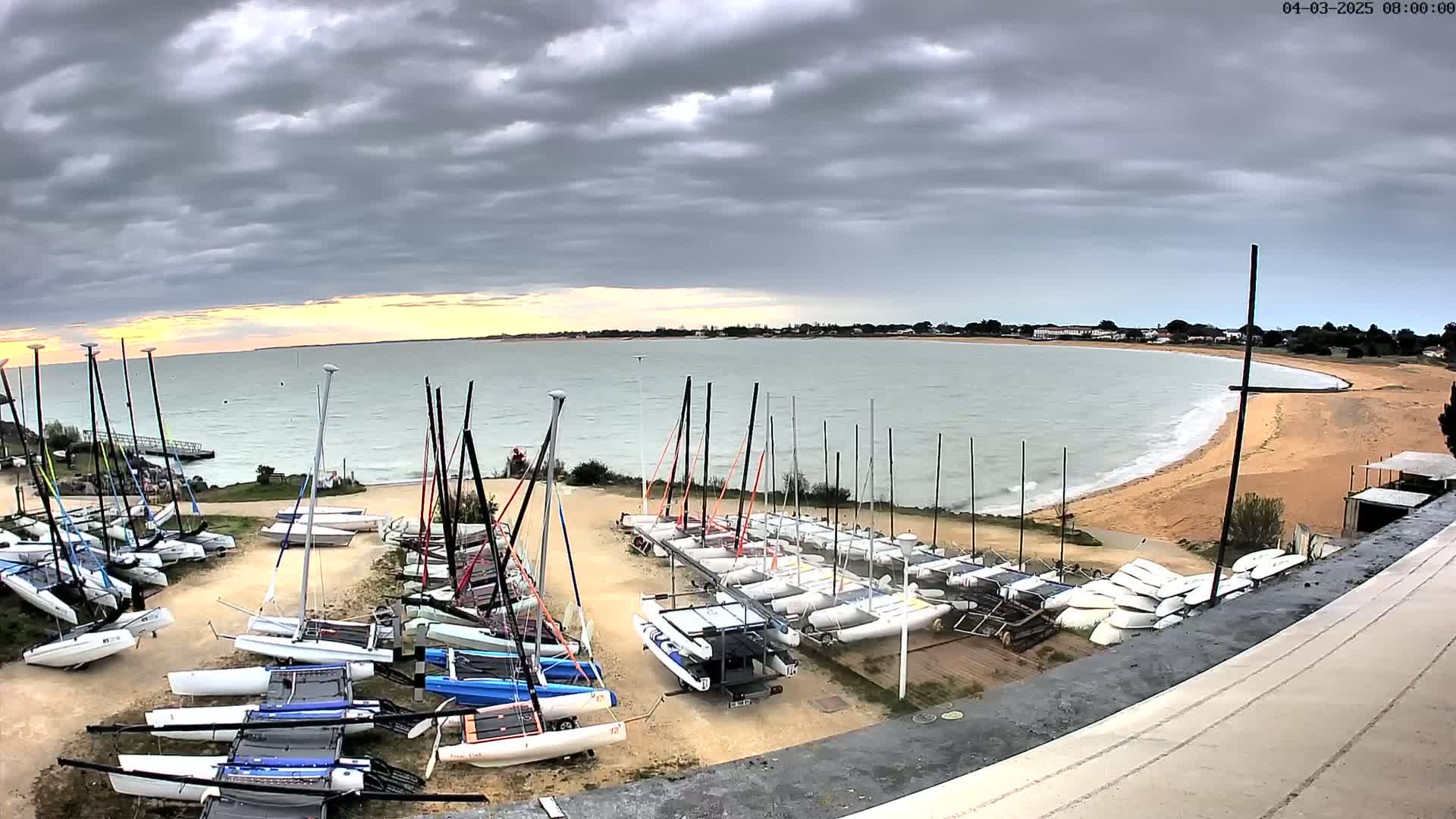Many sailboats are stored on a beach near a calm body of water under a cloudy sky.