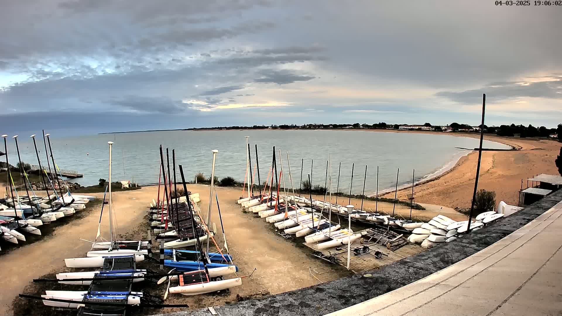 Numerous sailboats are docked on a sandy shore next to a calm body of water under a cloudy sky.