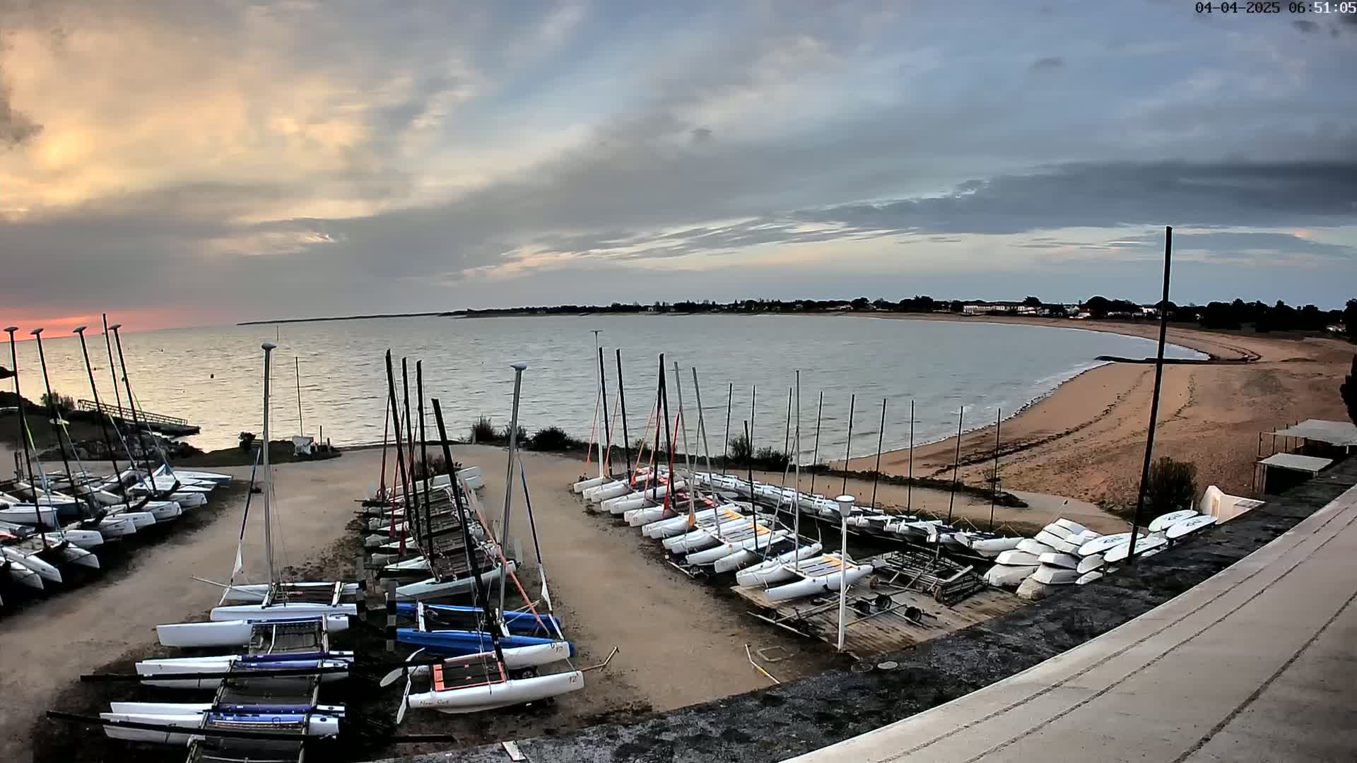 Many sailboats are stored on a sandy lot next to a calm body of water under a partly cloudy sky.
