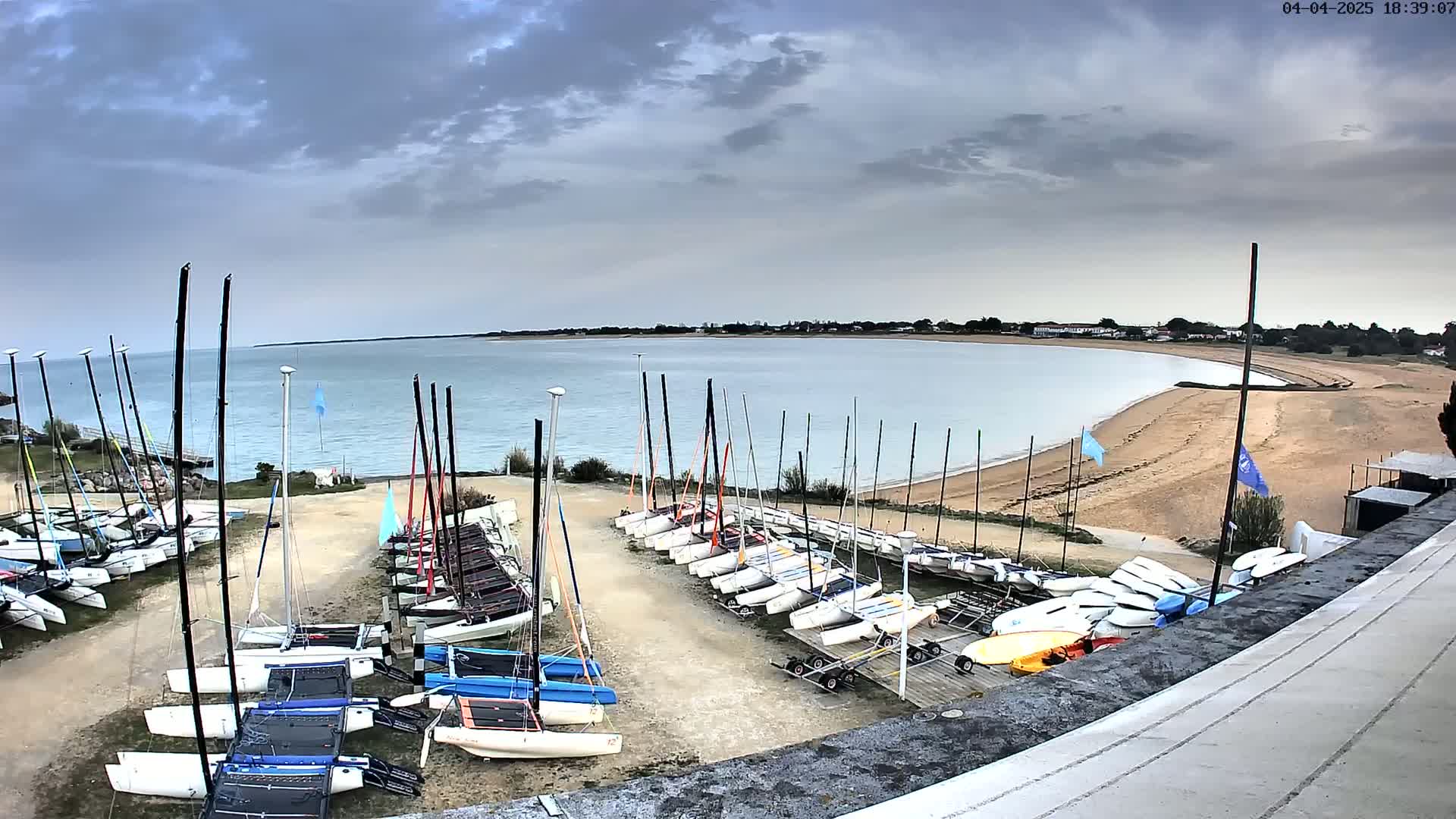 Numerous sailboats are docked on a sandy shore next to calm, light-blue water under a cloudy sky.