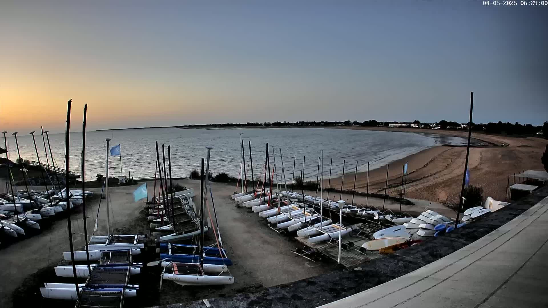 Many sailboats are docked at a calm, sandy beach during a sunrise with a clear sky.