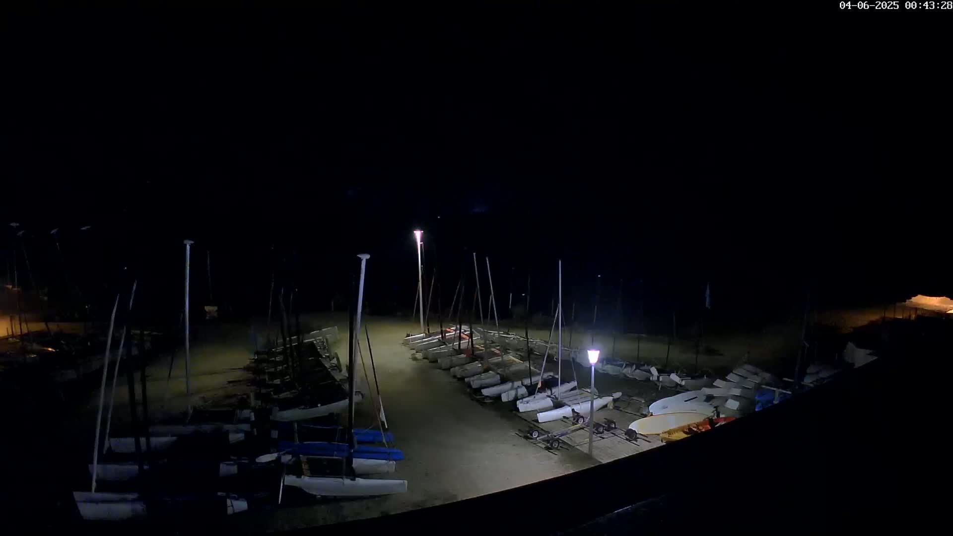 A nighttime, outdoor view of numerous small sailboats moored on a sandy area, illuminated by several lights.