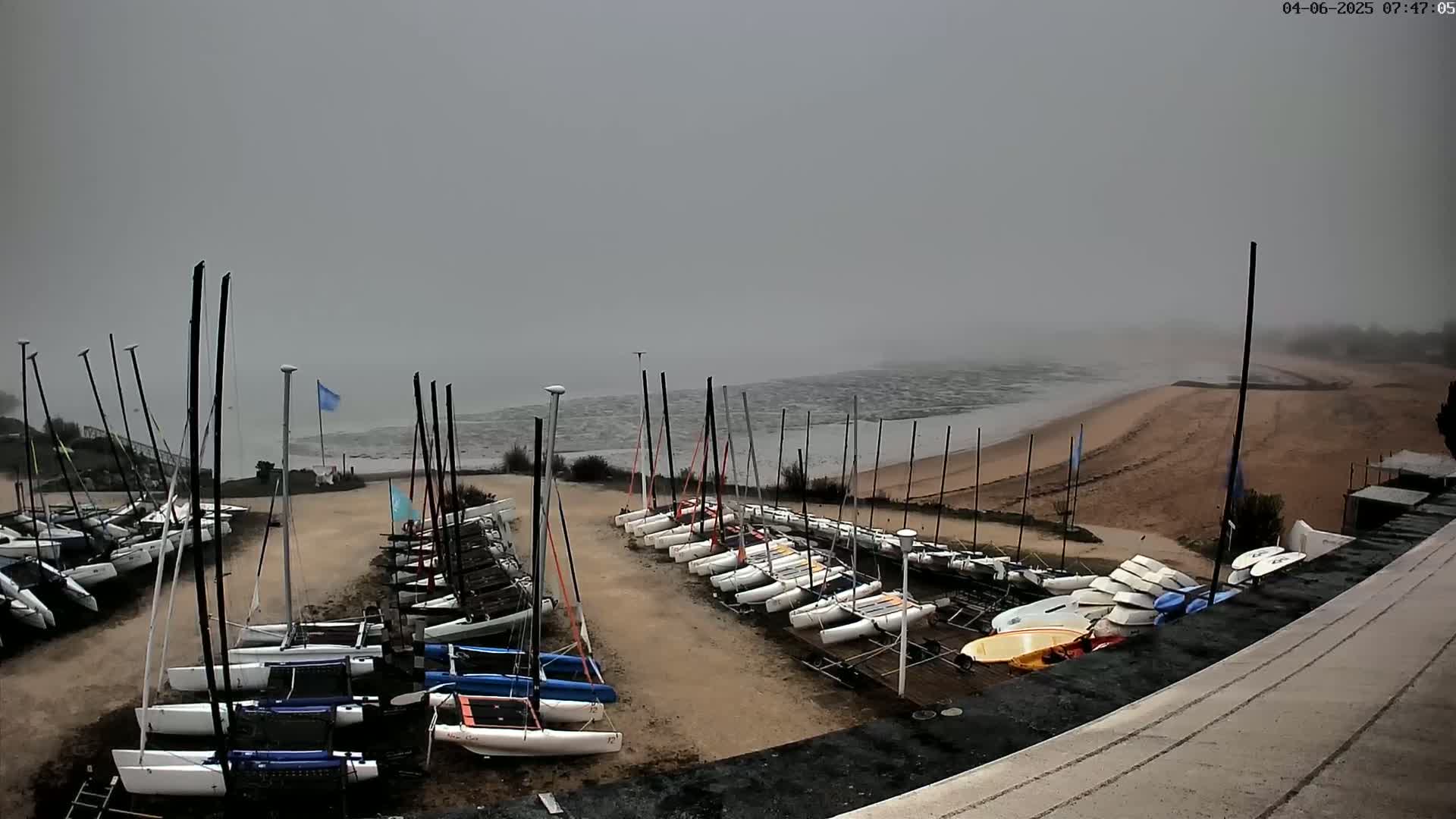 Numerous sailboats are parked on a sandy area next to a foggy ocean.