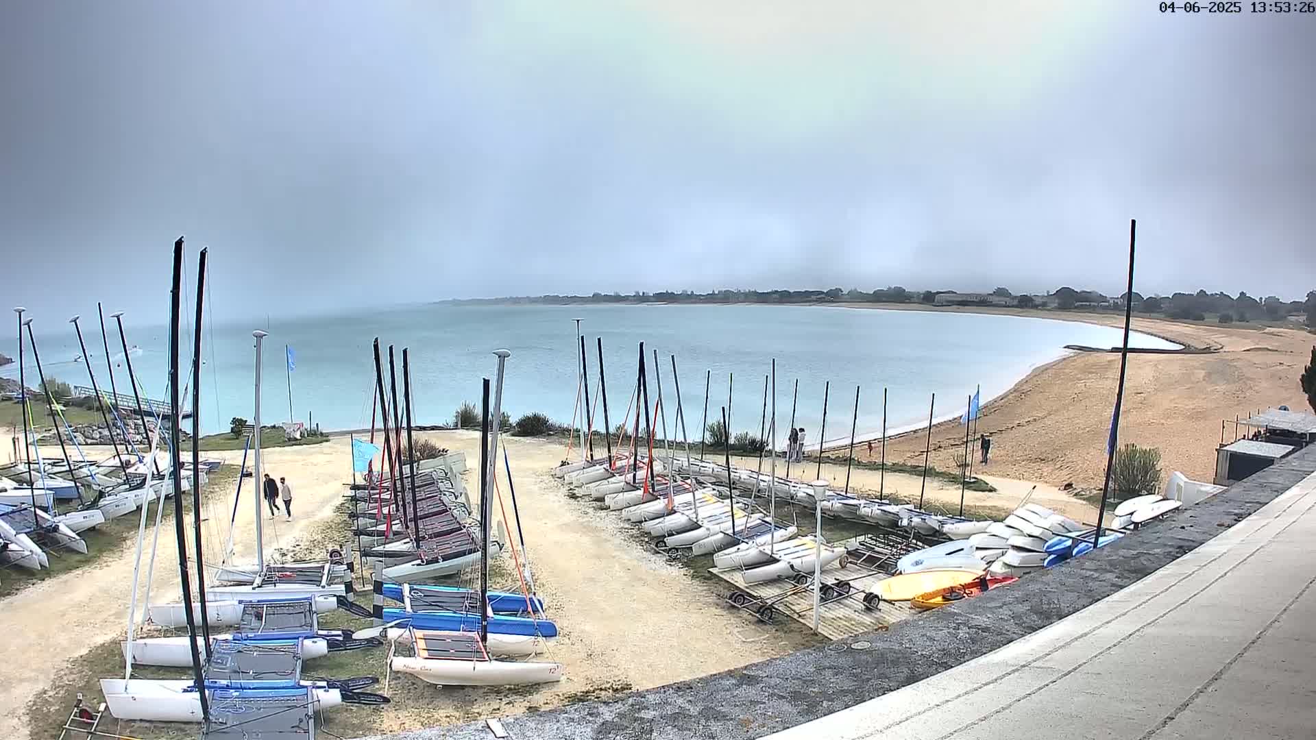 Numerous sailboats are stored on a sandy area next to a calm, light-blue body of water under an overcast sky.