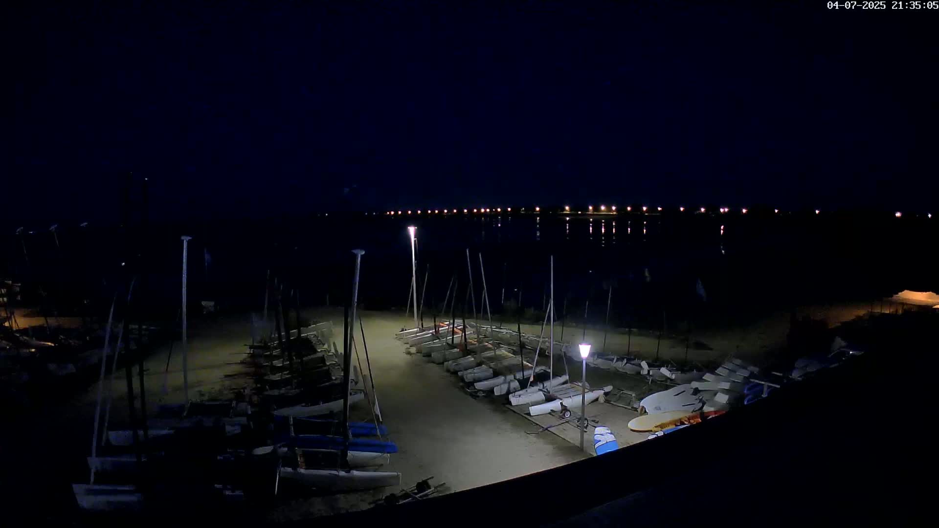 Many sailboats are parked on a sandy shore at night next to a calm body of water with distant lights.