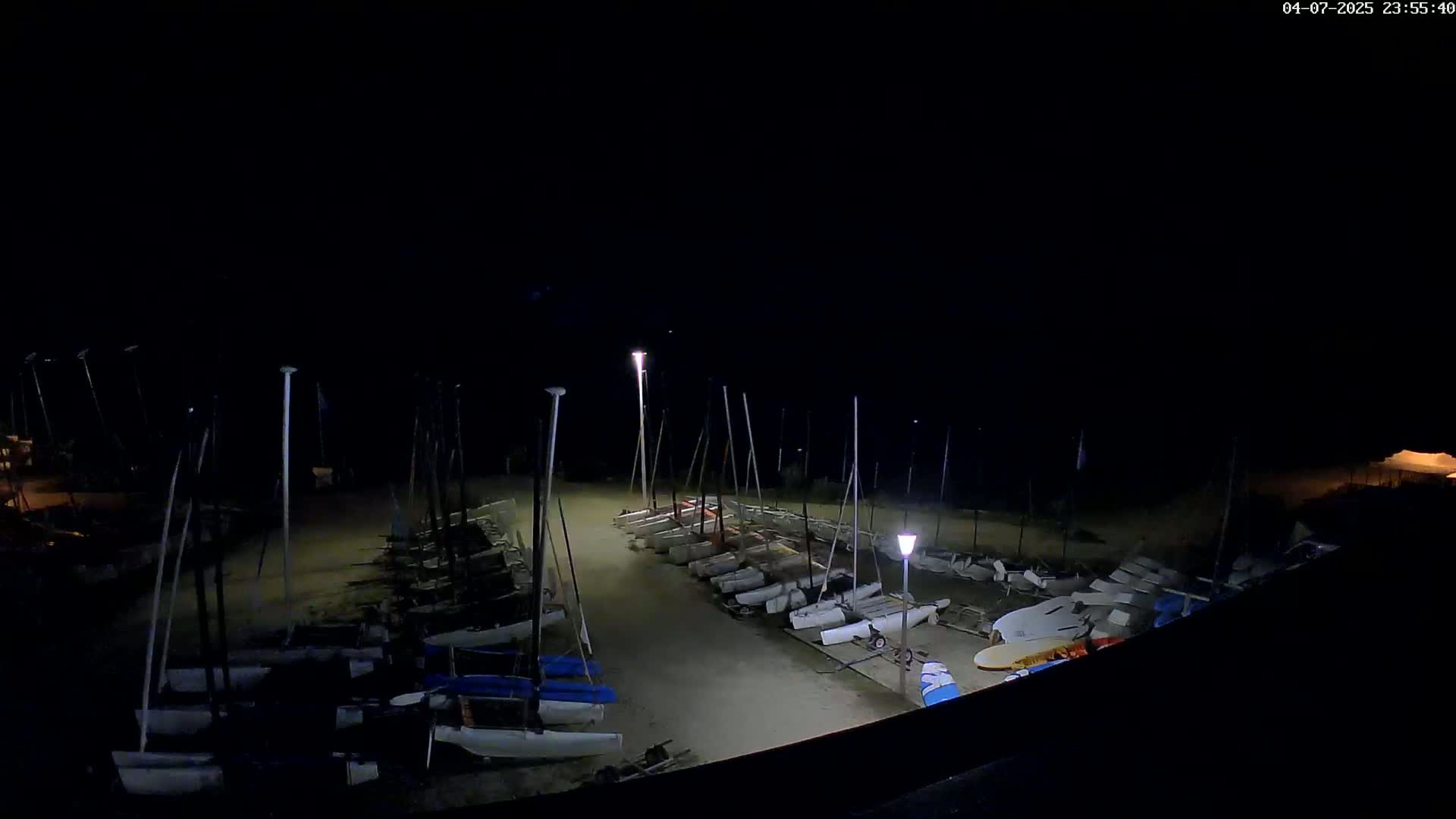 Numerous sailboats are docked in a sandy area at night under dim lighting.