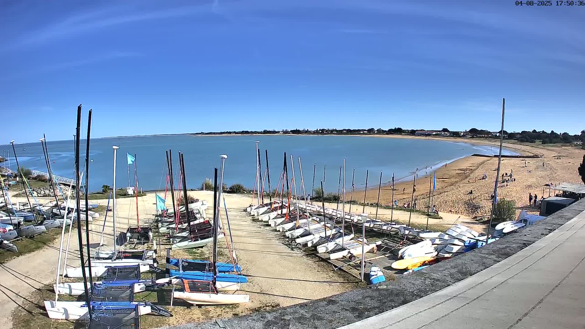 Many sailboats are stored on a sandy area next to a calm body of water with a sandy beach under a clear blue sky.