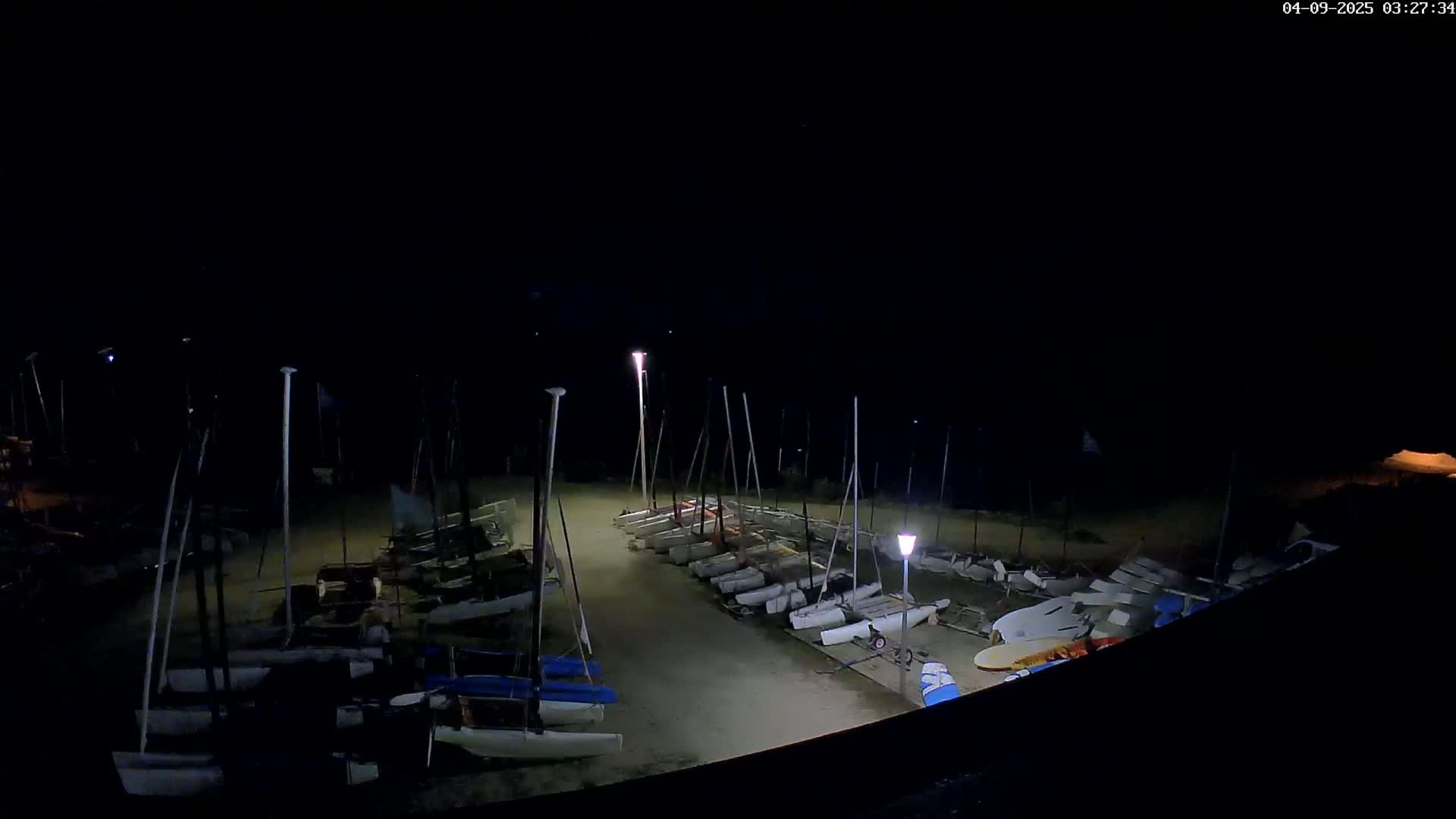 Numerous sailboats are docked at night on a sandy area, illuminated by several lights.