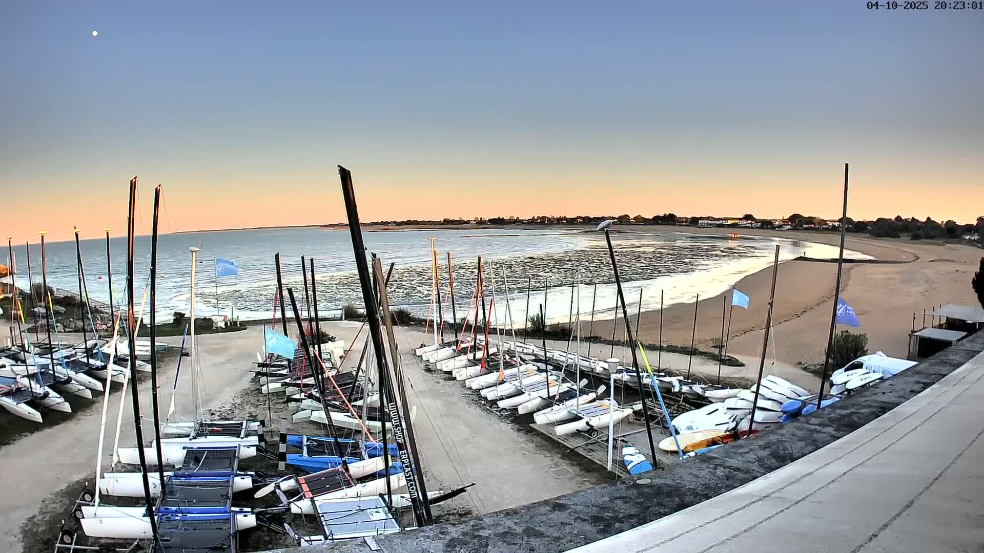 Many catamarans are parked on a sandy area next to a calm body of water at sunset.