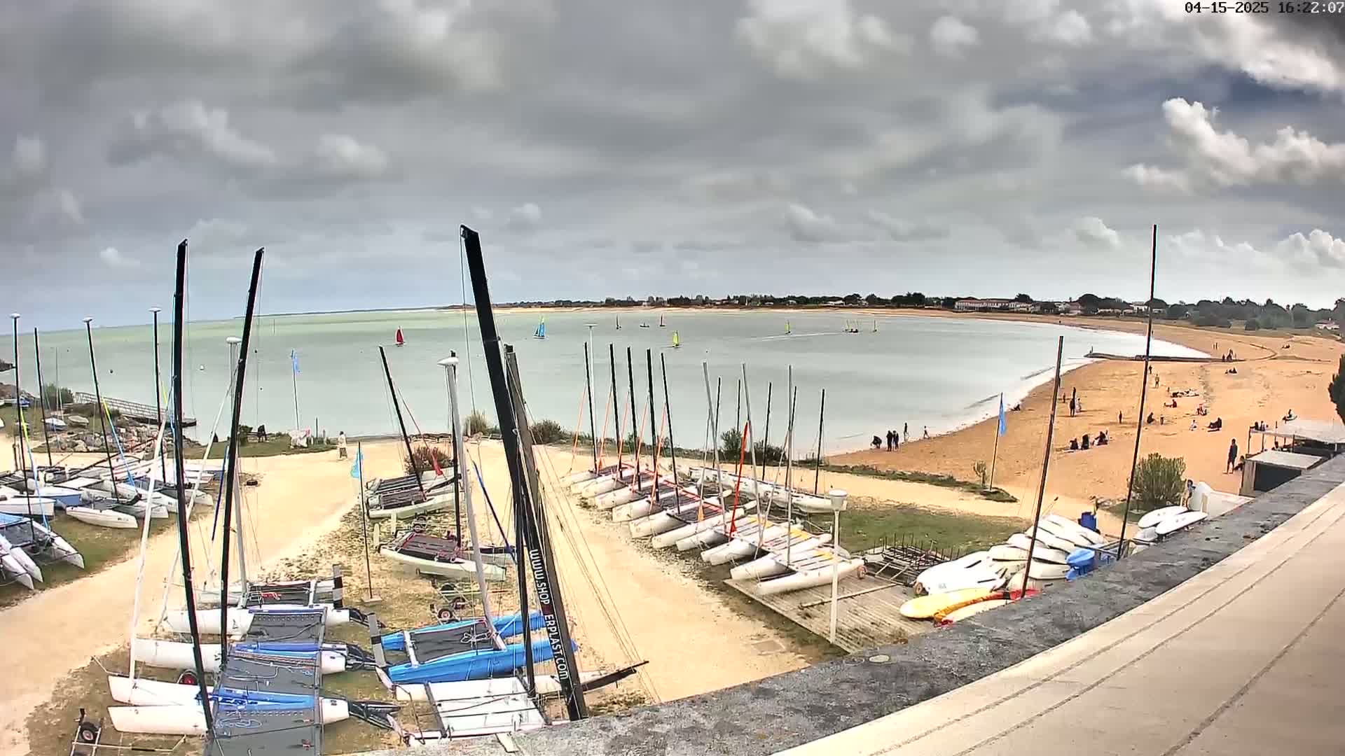 Numerous sailboats are docked near a sandy beach with people relaxing under a cloudy sky.