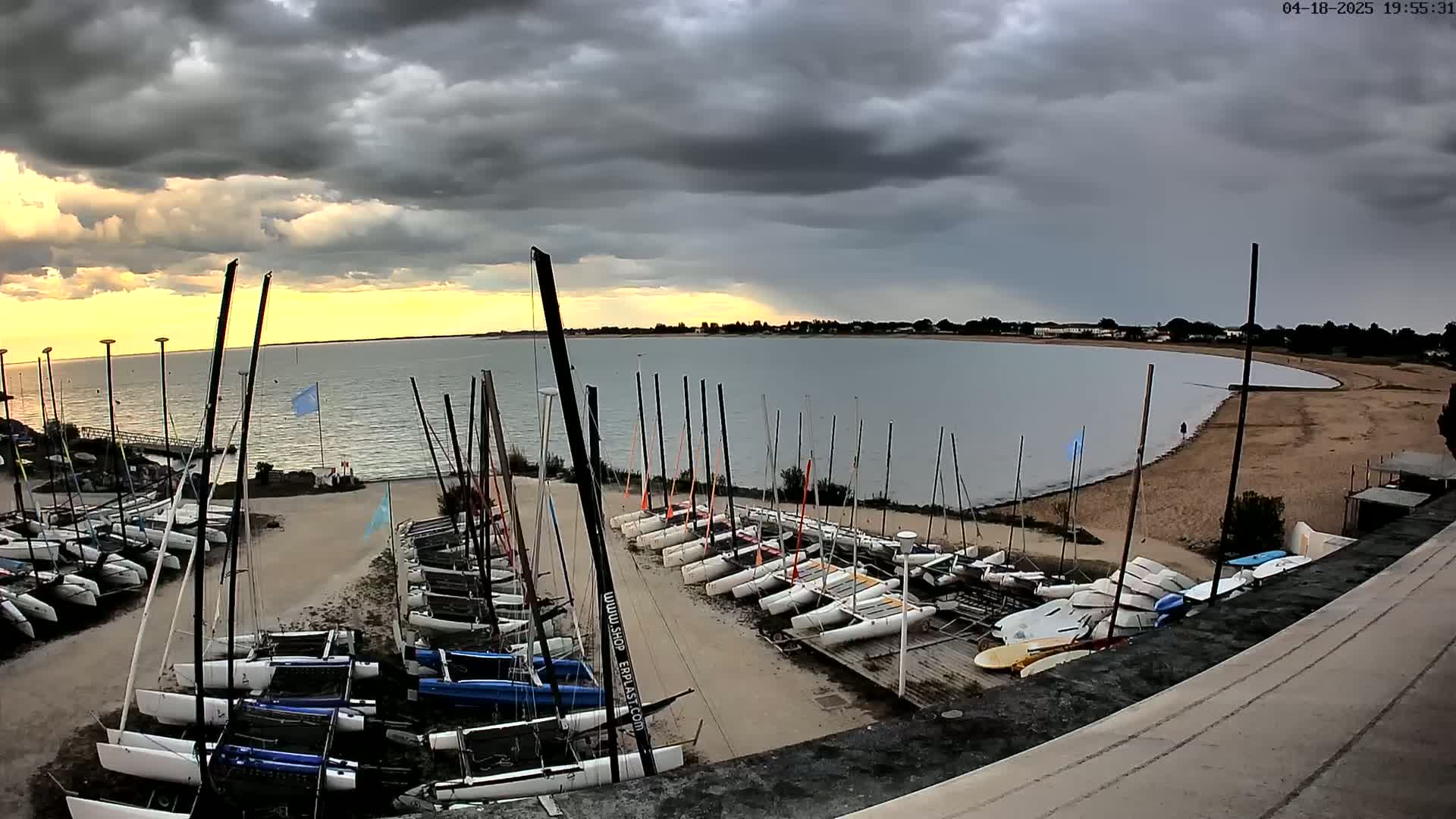 Many sailboats are docked at a sandy shore under a partly cloudy, stormy sky.