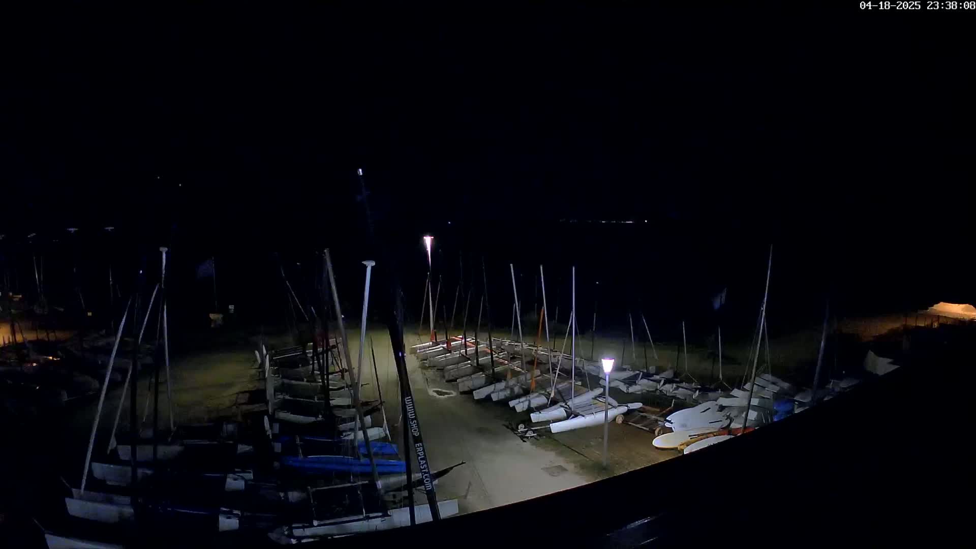 At night, numerous small sailboats are docked in a row along a shoreline under dim lighting.
