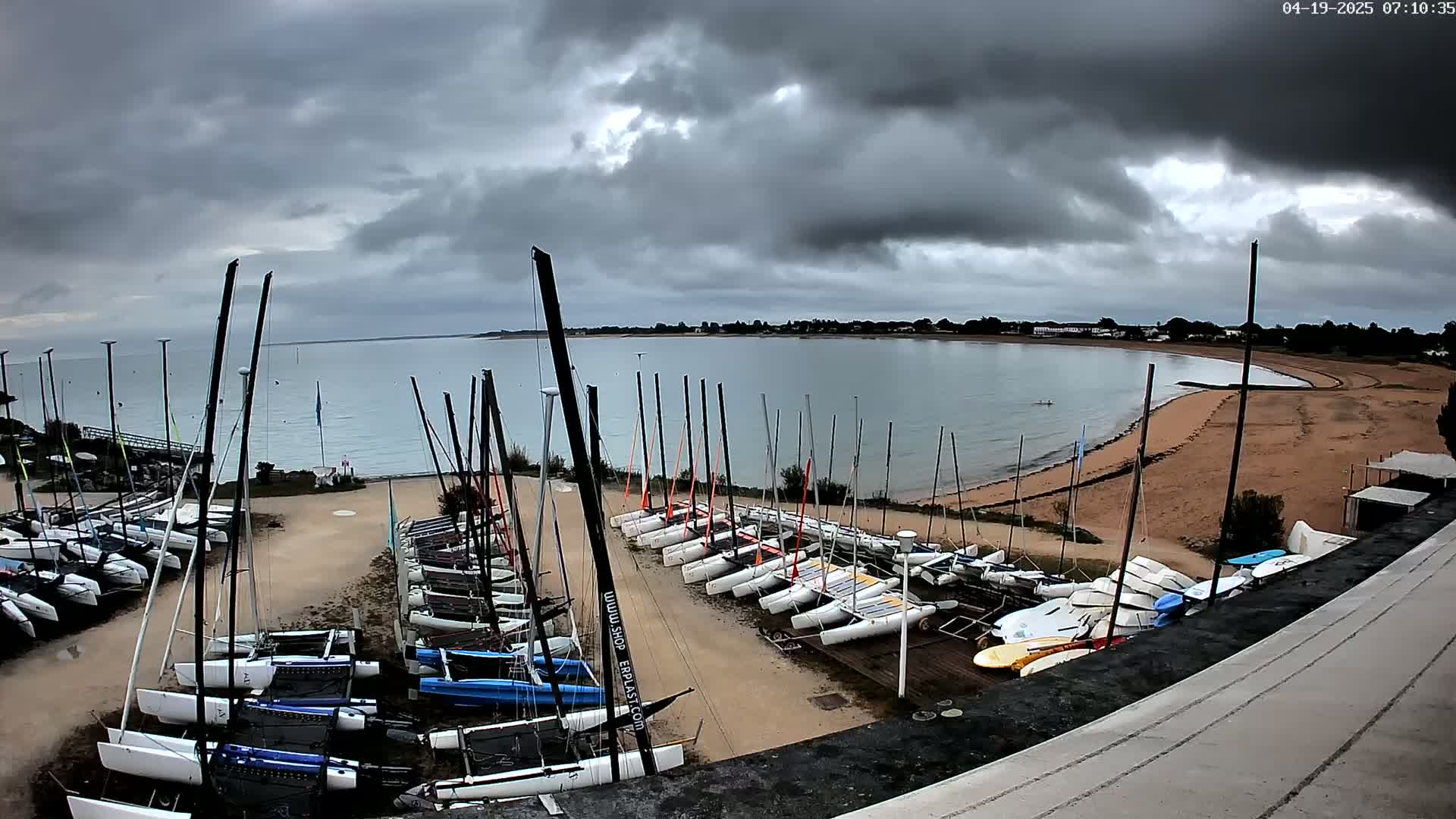 Many sailboats are docked on a sandy shore next to calm water under a cloudy sky.