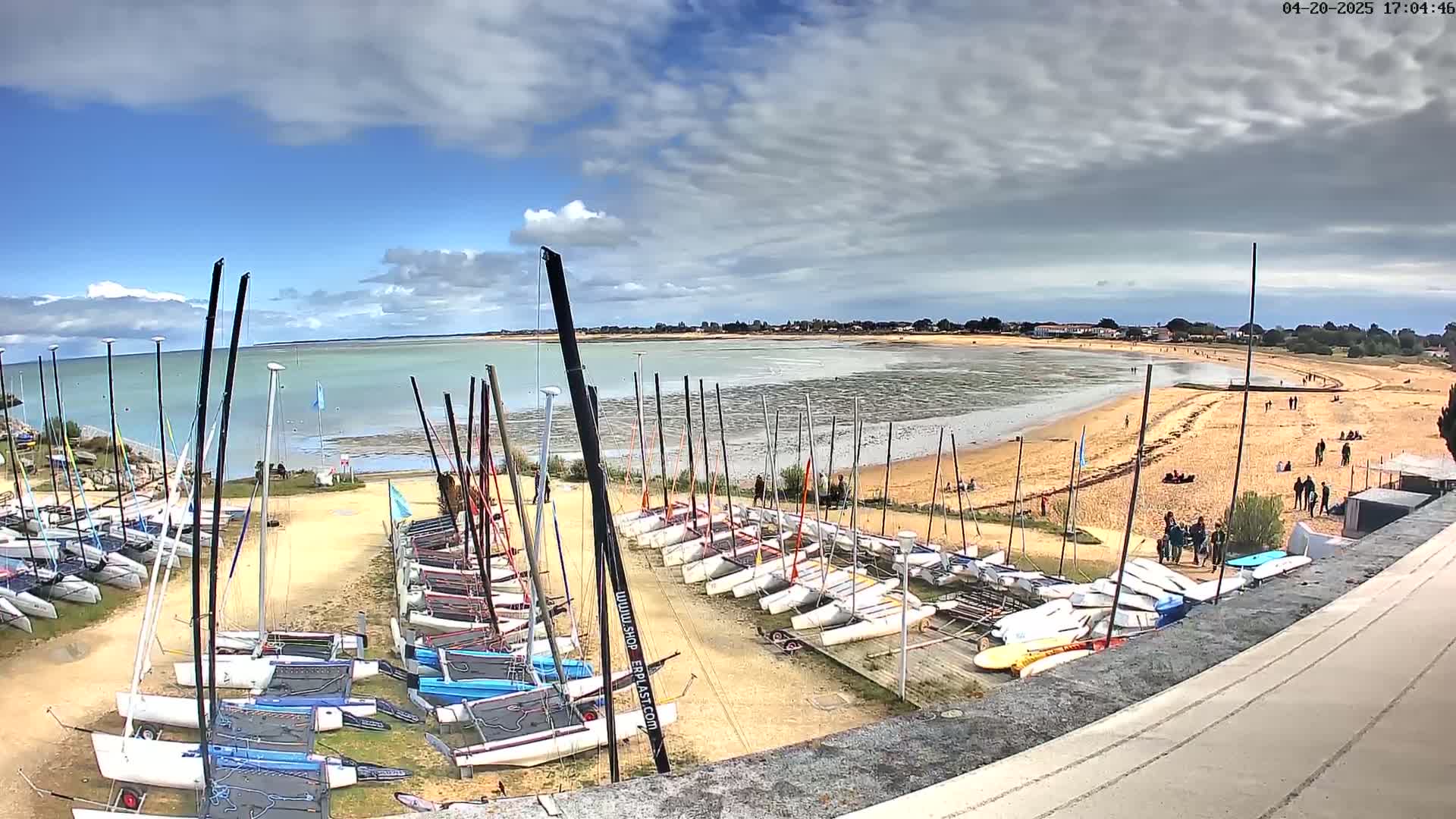 Numerous sailboats are parked on a sandy beach next to calm, shallow water under a partly cloudy sky.
