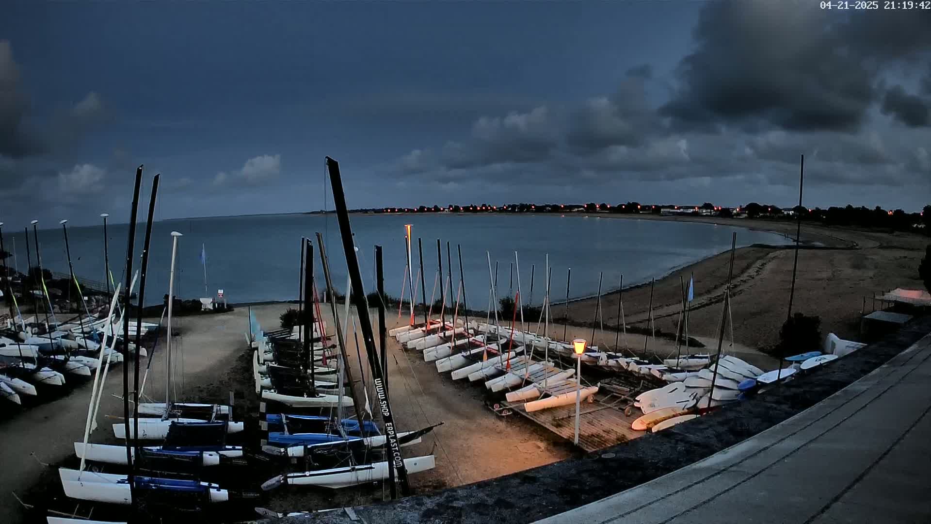 Many sailboats are docked on a sandy shore at dusk under a cloudy sky.