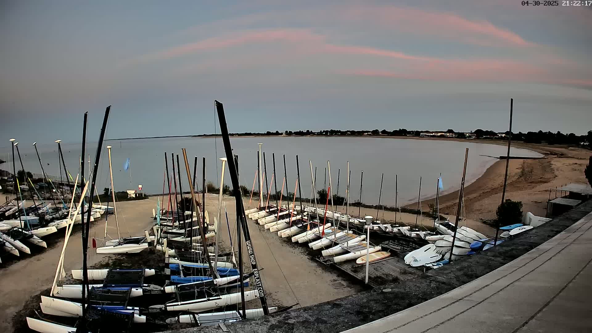 Numerous sailboats are docked on a sandy shore next to calm water under a pale pink and gray sky.