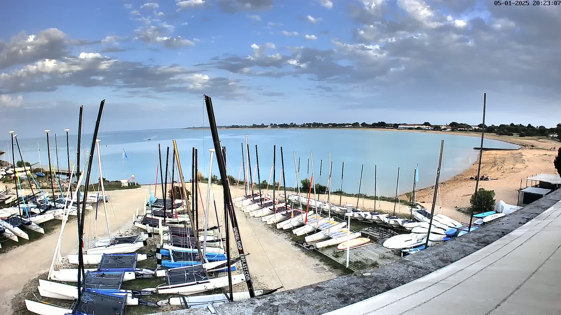 Numerous sailboats are docked on a sandy shore next to a calm, light-blue body of water under a partly cloudy sky.