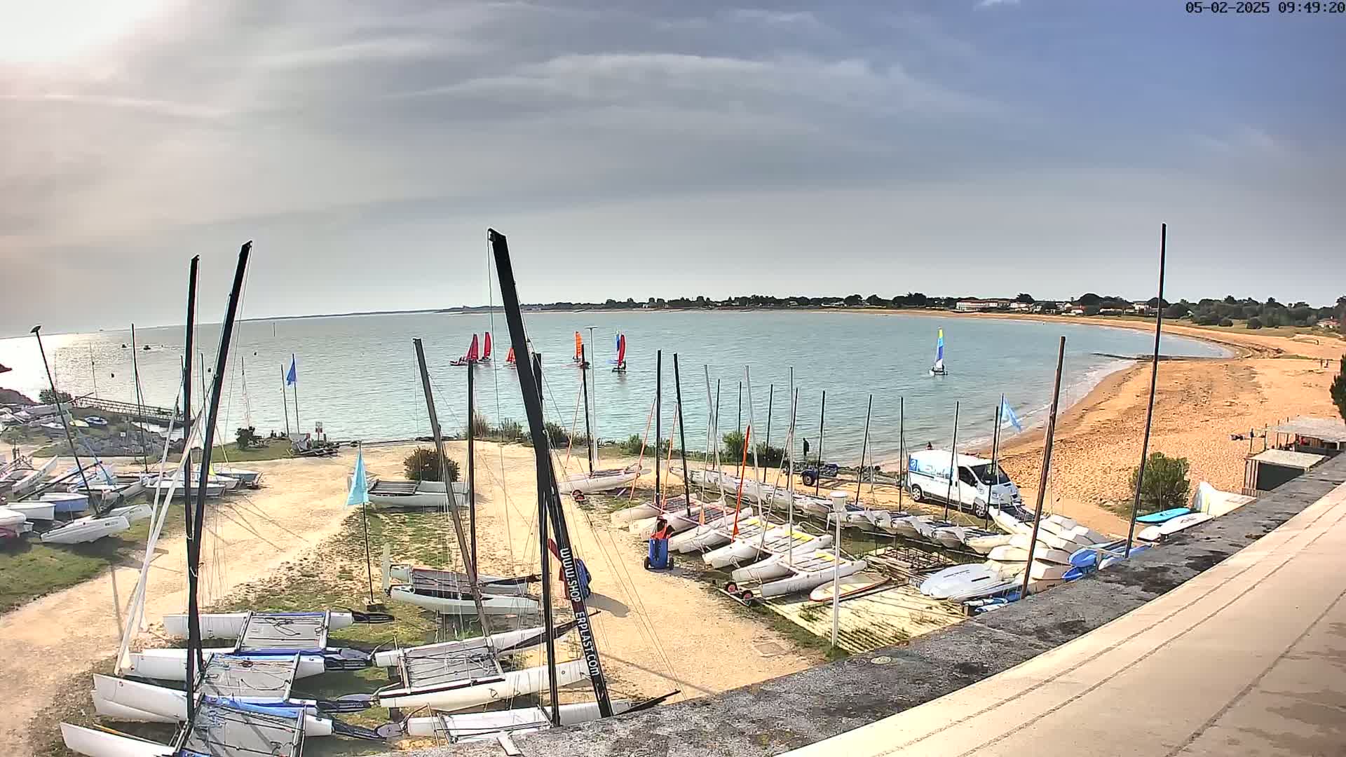 Many sailboats are docked on a sandy shore next to a calm body of water under a partly cloudy sky.