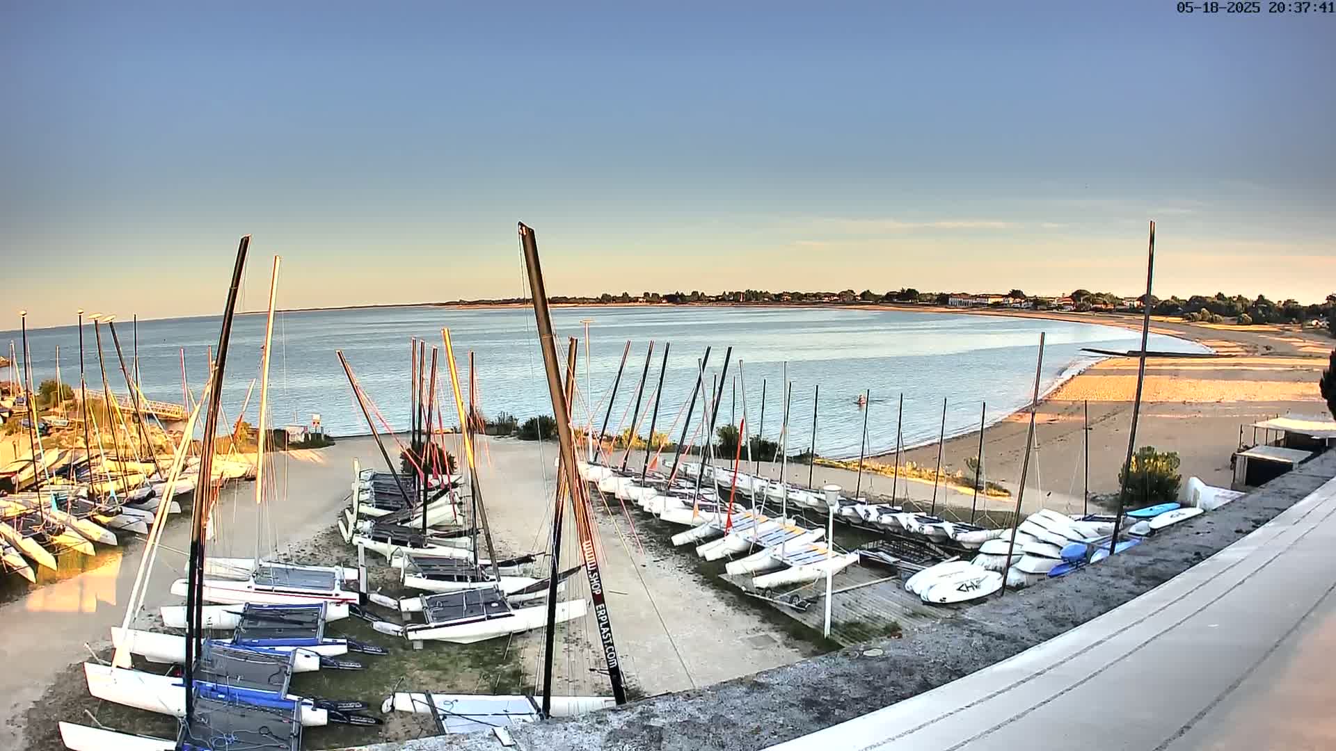 Numerous sailboats are docked on a sandy shore next to calm, clear water under a mostly clear sky at sunset.