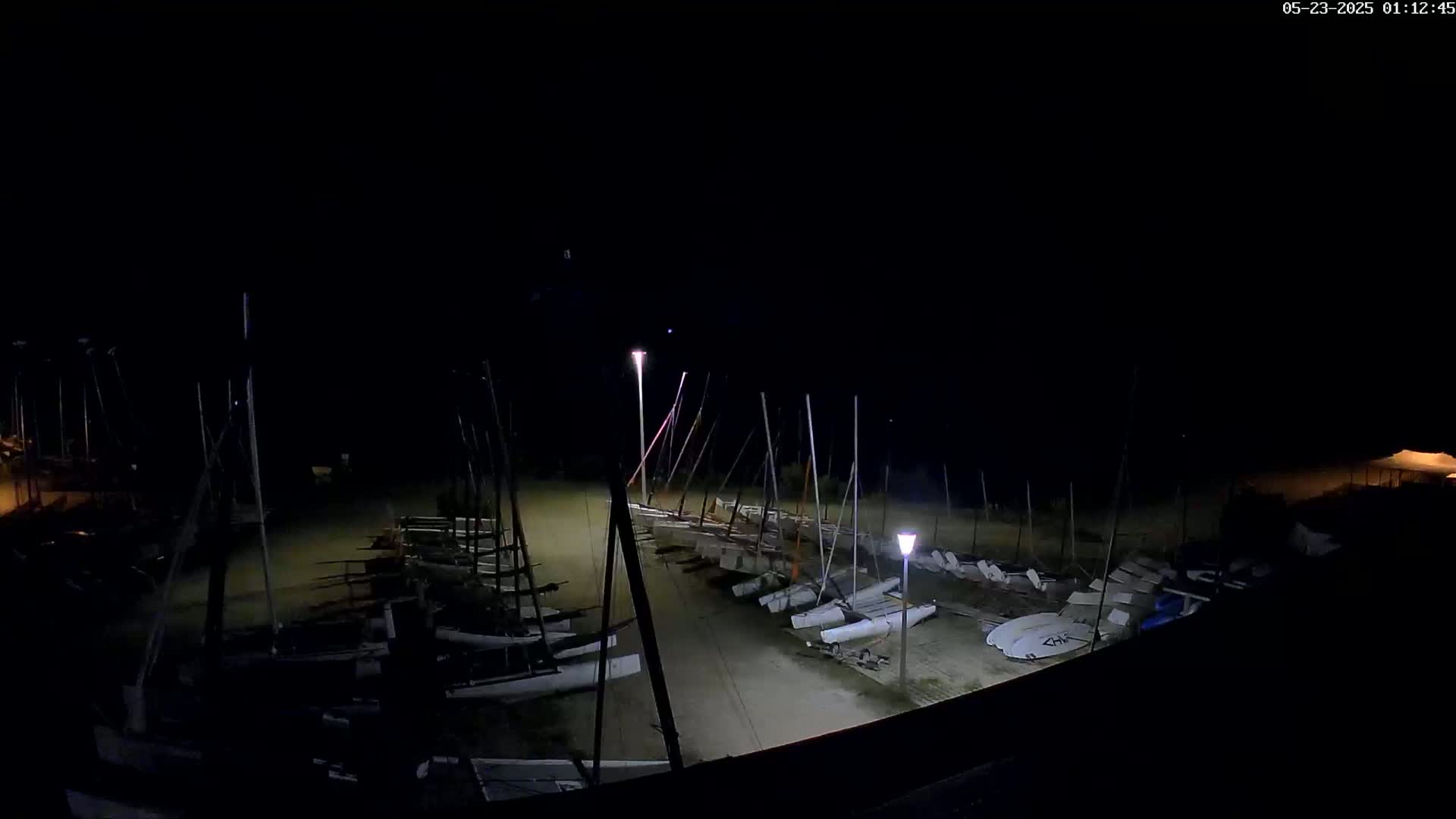 A nighttime, high-angle view shows numerous sailboats beached on a sandy area, illuminated by several lights.