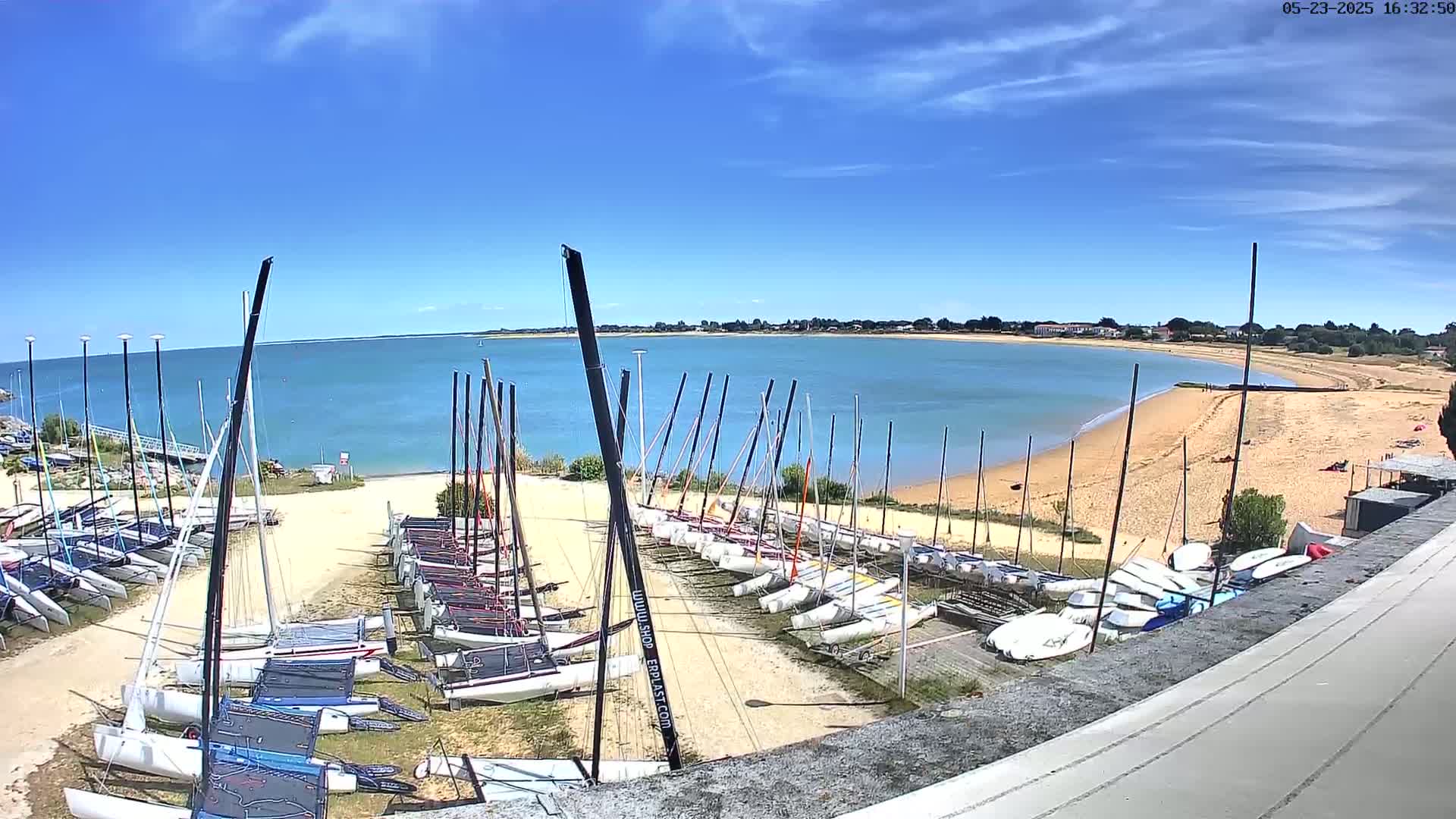 Numerous sailboats are stored on a sandy area next to a calm body of water under a bright, sunny sky.