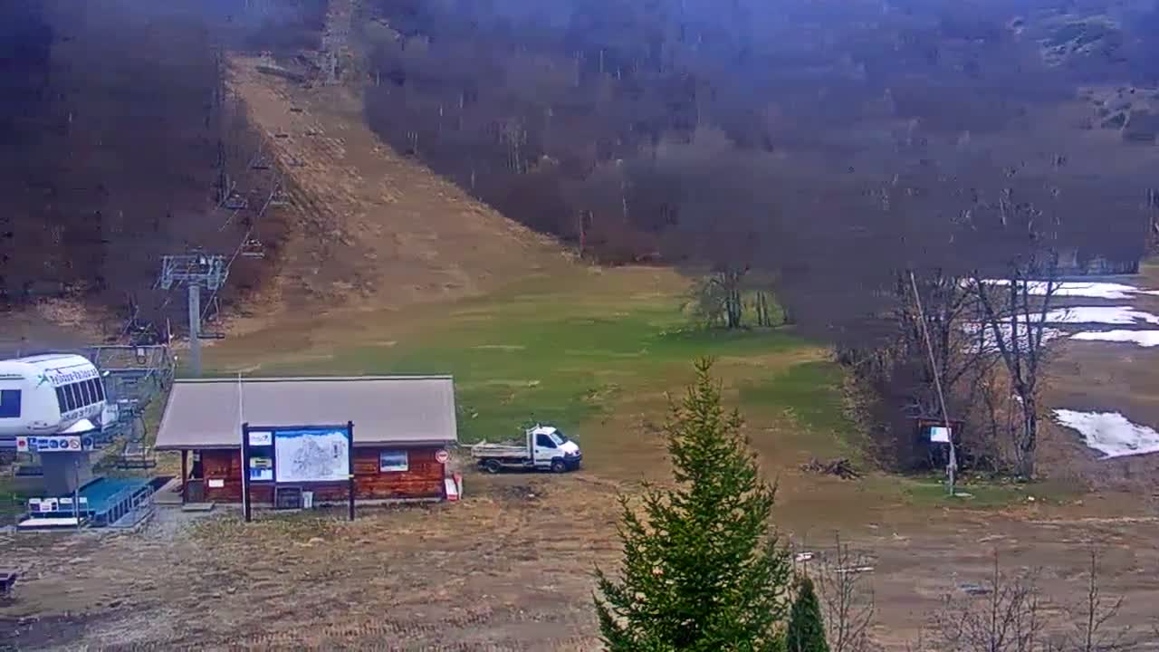 A ski lift base station with a small building and a white truck is situated on a mostly brown, grassy hill under an overcast sky.