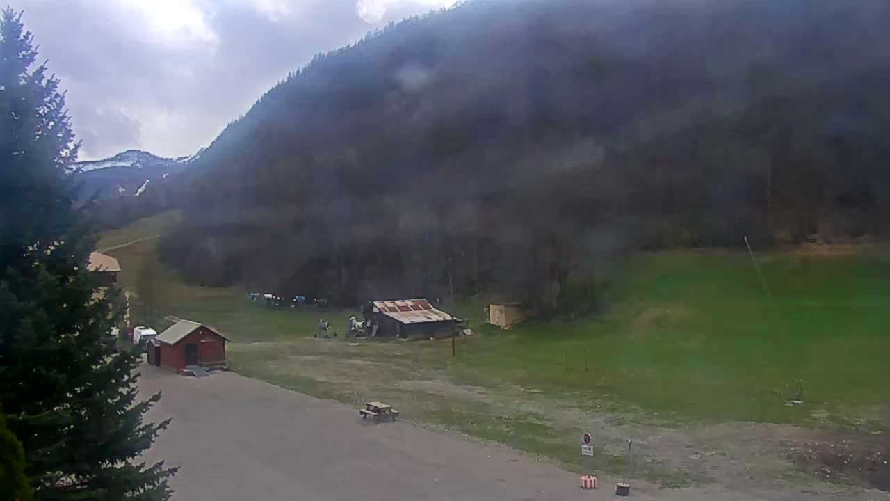 A mostly cloudy day reveals a mountainous landscape with a small, rusty shed, some outbuildings, and a few animals in a grassy field near a paved area.