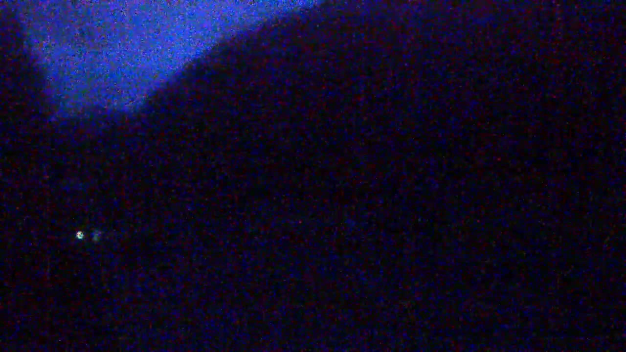 A dark, nighttime scene shows a blurry, indistinct landscape under a mostly clear, dark blue sky, with a single, small light visible in the distance.