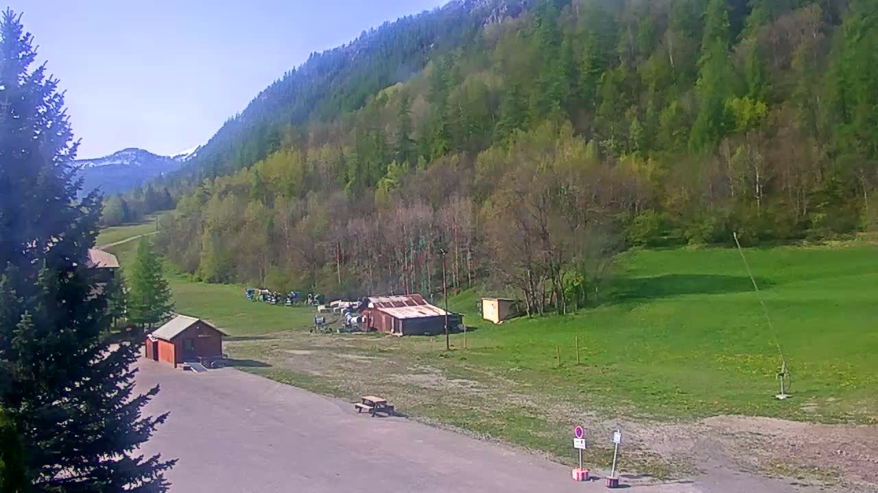 A sunny view of a grassy field and a hillside covered in lush green trees, with a few small buildings and a picnic table in the foreground.