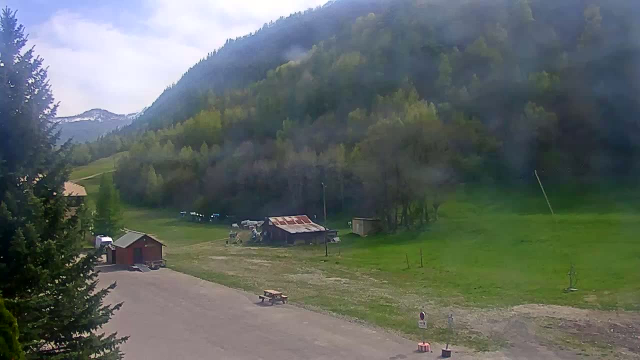 A hazy view shows a mountainous area with green fields, a few small buildings, and a paved area near evergreen trees under a partly cloudy sky.