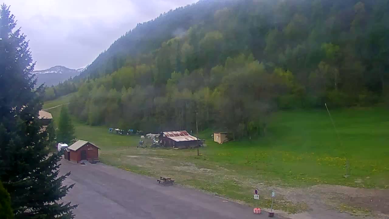A paved area with a small building and a few picnic tables sits before a grassy field, which is nestled at the base of a misty, verdant mountainside under an overcast sky.