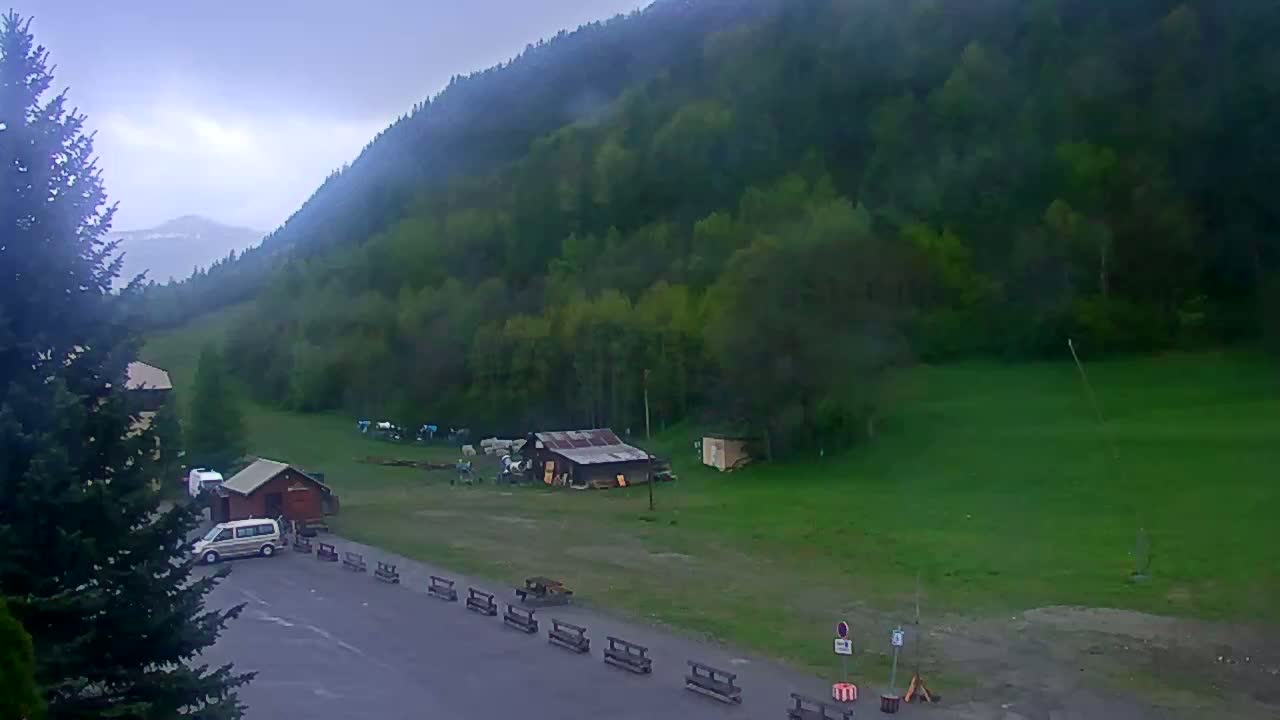 A cloudy day shows a mountainside covered in lush green trees, with a small parking lot containing several vehicles near some small buildings and benches.