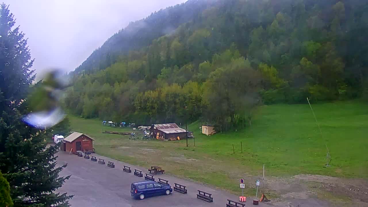 A rain-covered view shows a dark blue car parked in a lot next to a small building, with a grassy field, some small sheds, and a large, lush green hillside beyond.