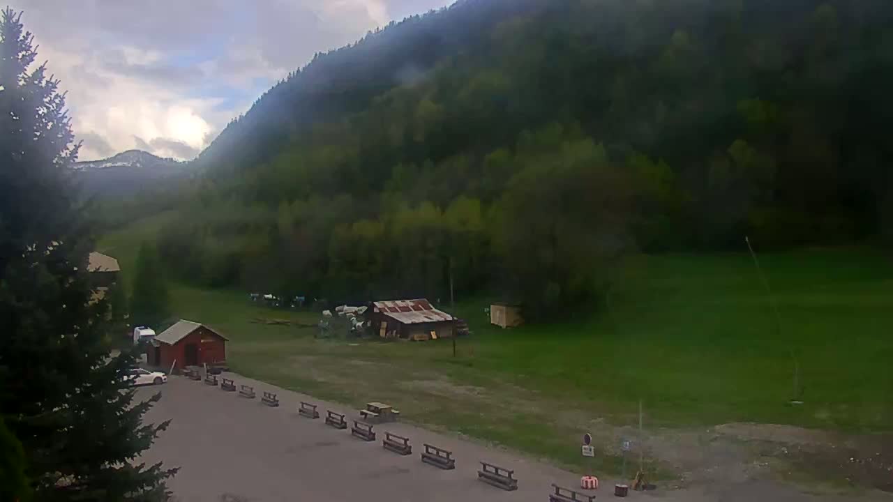 A cloudy day reveals a mountainside covered in lush green trees, with a small parking area below containing a few vehicles and several benches.