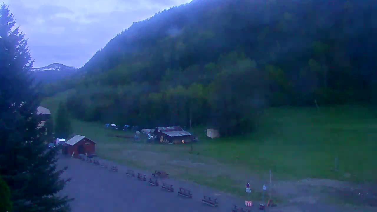 A twilight view shows a grassy area with a few small buildings near a large, dark green mountain under a cloudy sky.