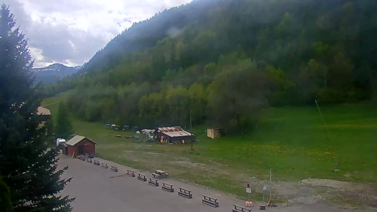A cloudy day reveals a mountainside covered in lush green trees, a small parking area with picnic benches, and a few small buildings.