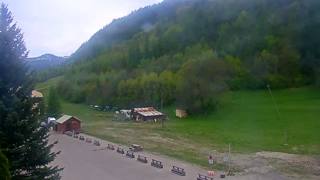 A mostly cloudy day reveals a mountainside covered in lush green trees, a small building, and a paved area with benches.