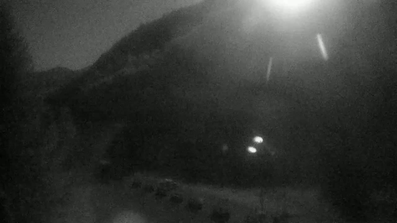 A nighttime view of a dark mountain silhouetted against a bright sky, with several indistinct vehicles parked below and lights visible in the distance.